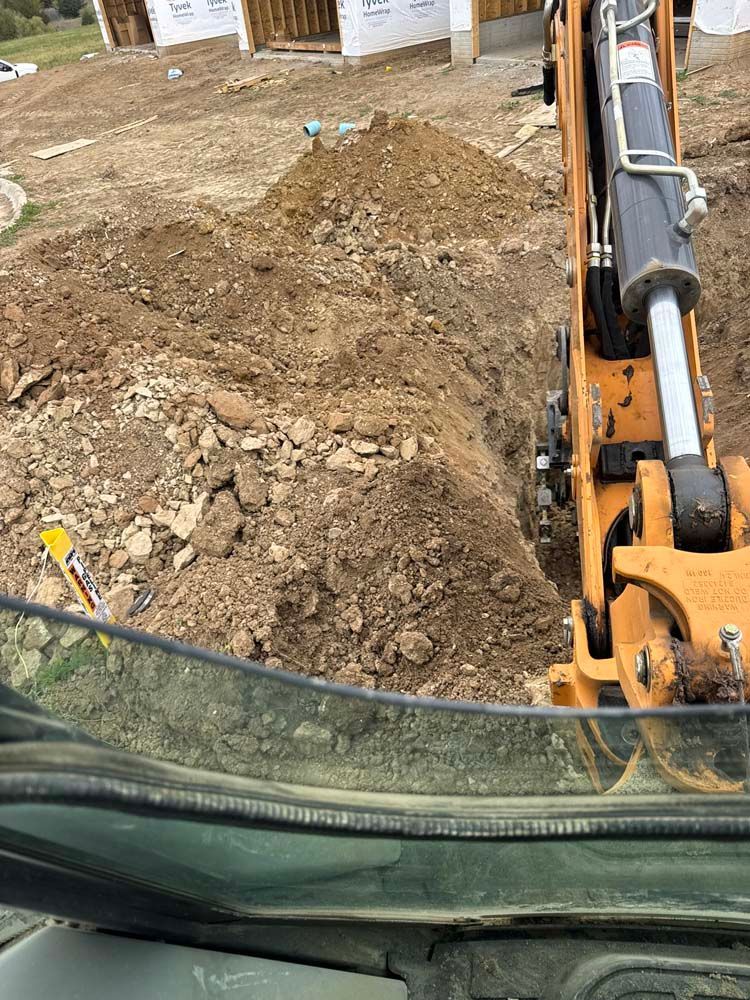Excavator digging a trench on a construction site. Dirt pile and new house under construction visible.