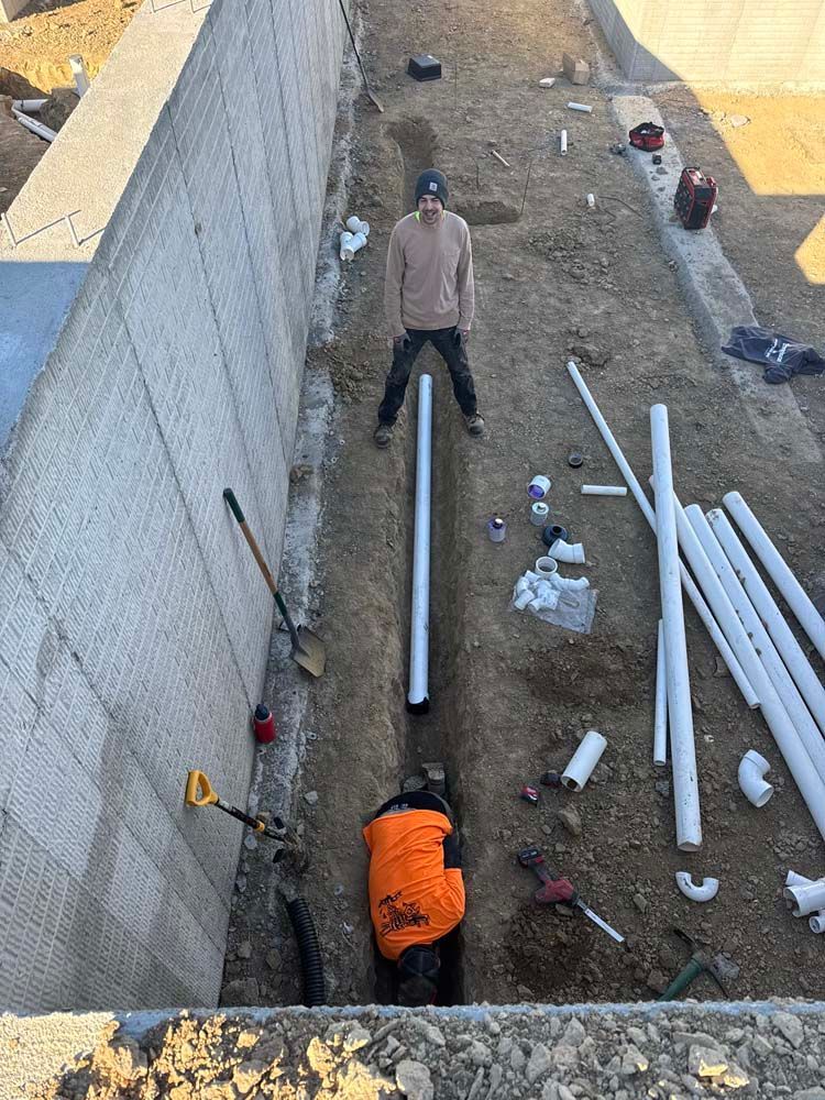 Two people installing pipes in a trench next to a concrete wall. Tools and materials are scattered around.