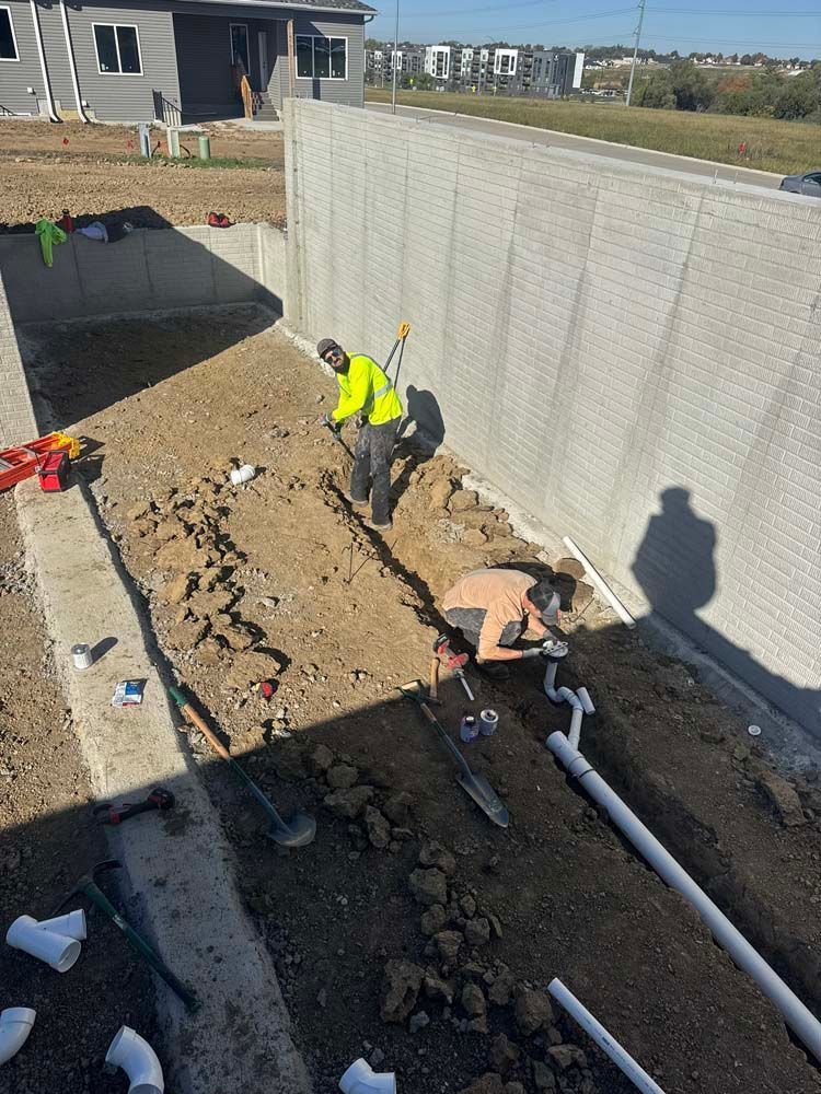 Construction workers laying pipes in a trench near a concrete wall. One digs with a shovel.