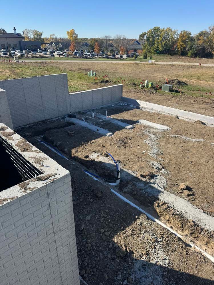 Construction site: concrete foundation with exposed pipes and soil. Background features buildings, trees, and clear sky.