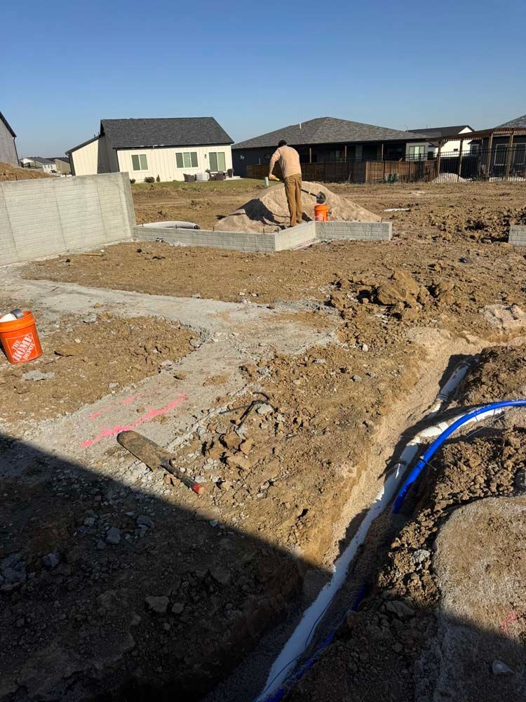 Construction site with a person working on a foundation, blue pipes visible, and residential houses in the background.