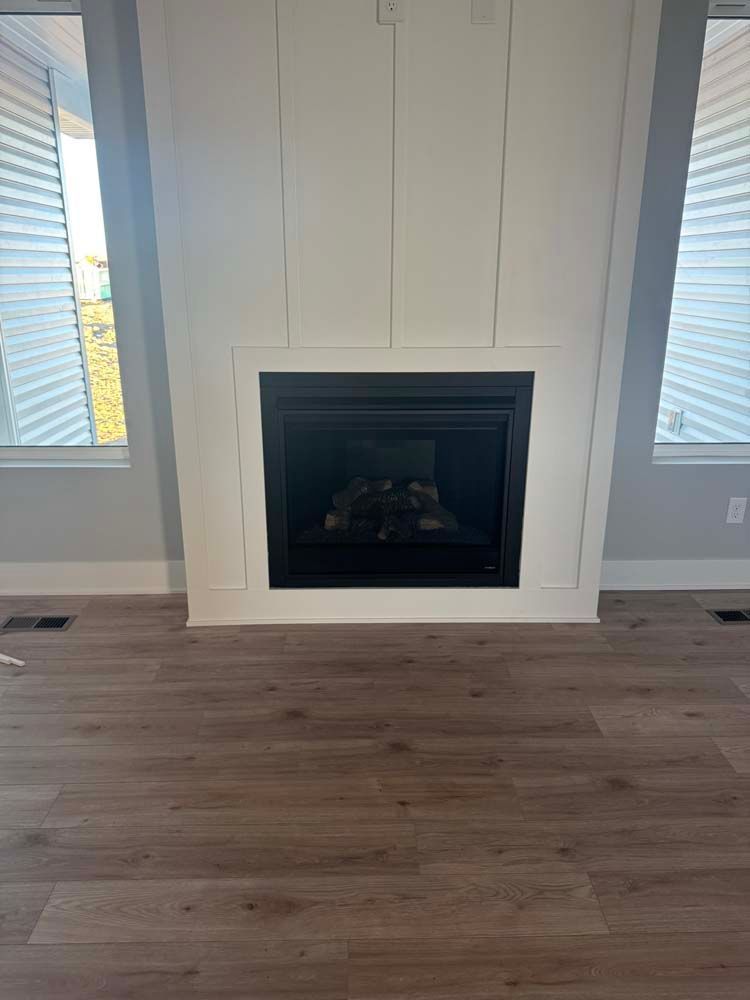 White fireplace built into a wall with paneling, flanked by windows with blinds, and hardwood floor.