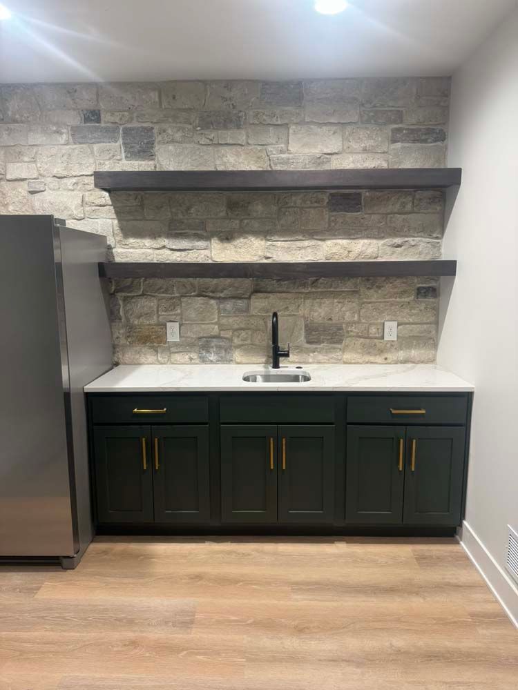 Dark green bar cabinets with gold hardware, white countertop, stone backsplash, shelves, and black faucet.