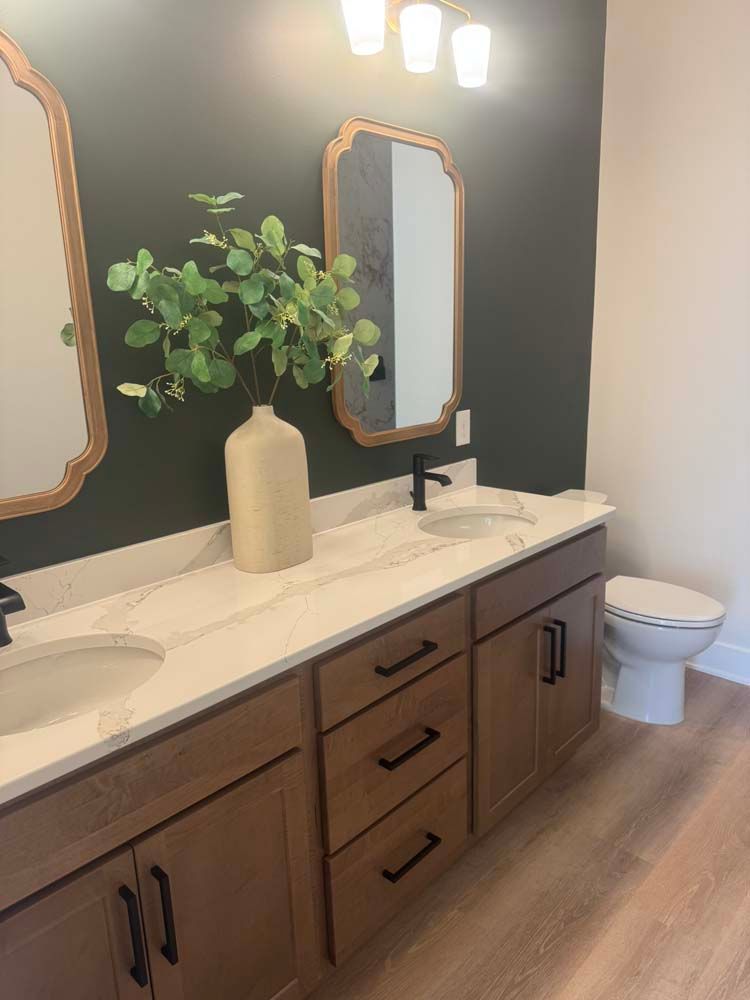 Bathroom with dark green wall, light wood vanity, white countertop, and gold-framed mirrors.