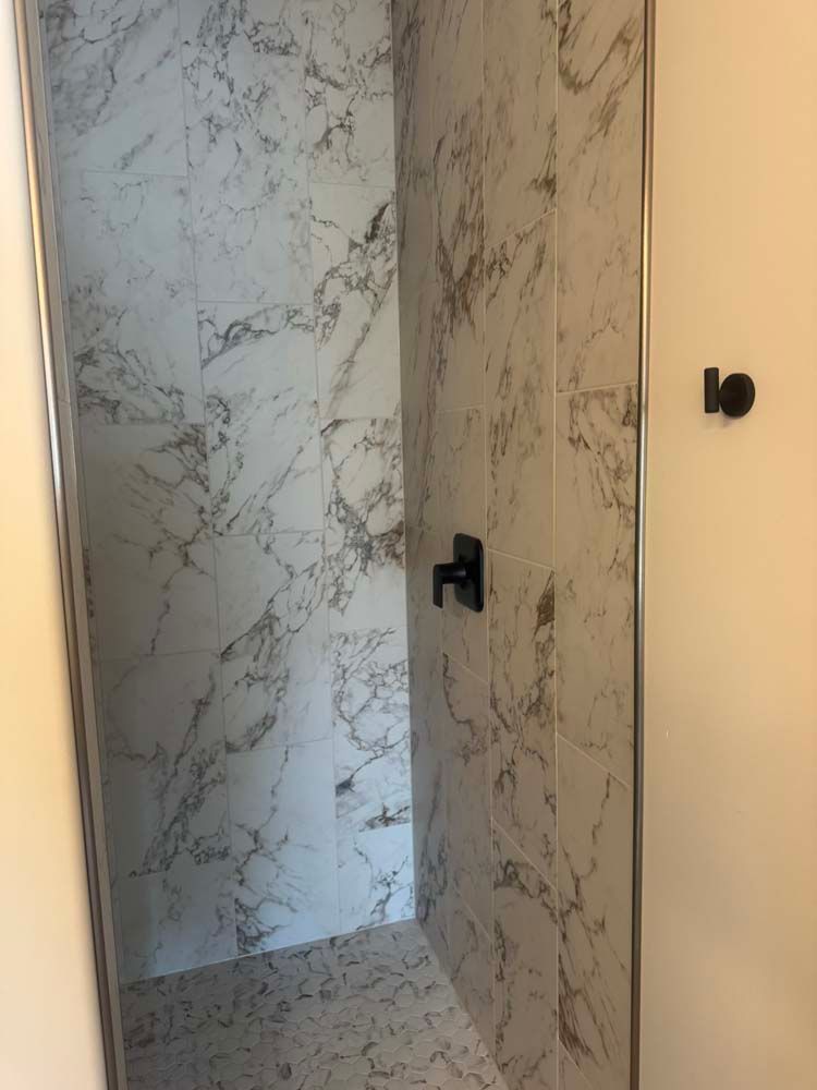 Marble-tiled shower with a black faucet. The shower has a clear glass door. A black hook is on the adjacent wall.