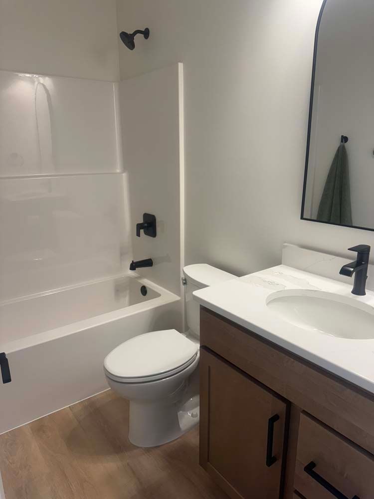 Modern bathroom with white tub, toilet, and sink, brown vanity, black fixtures, and light wood-look flooring.