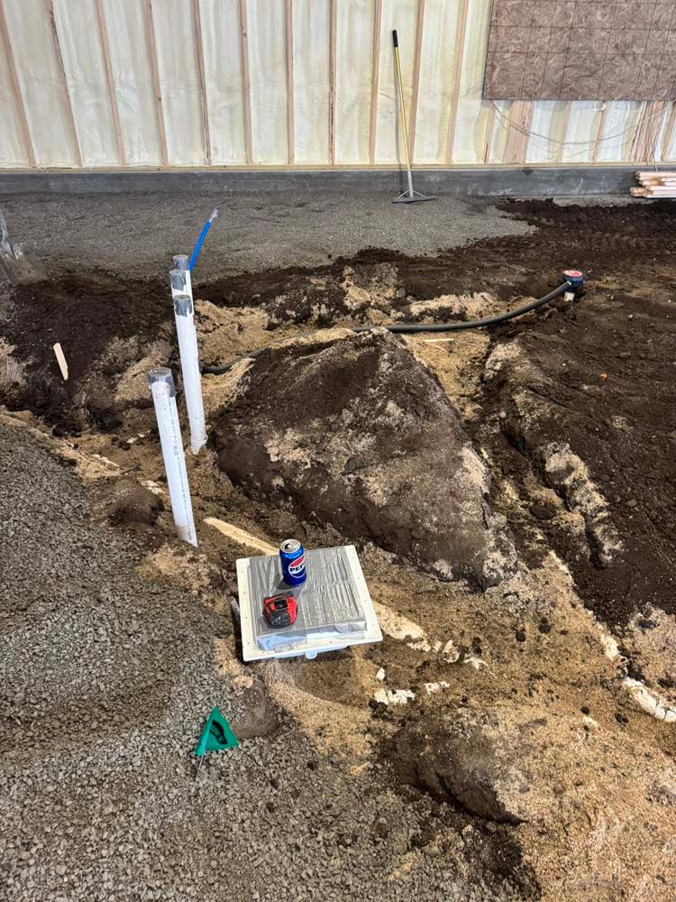 Construction site with plumbing pipes emerging from the dirt floor; tools and debris visible.