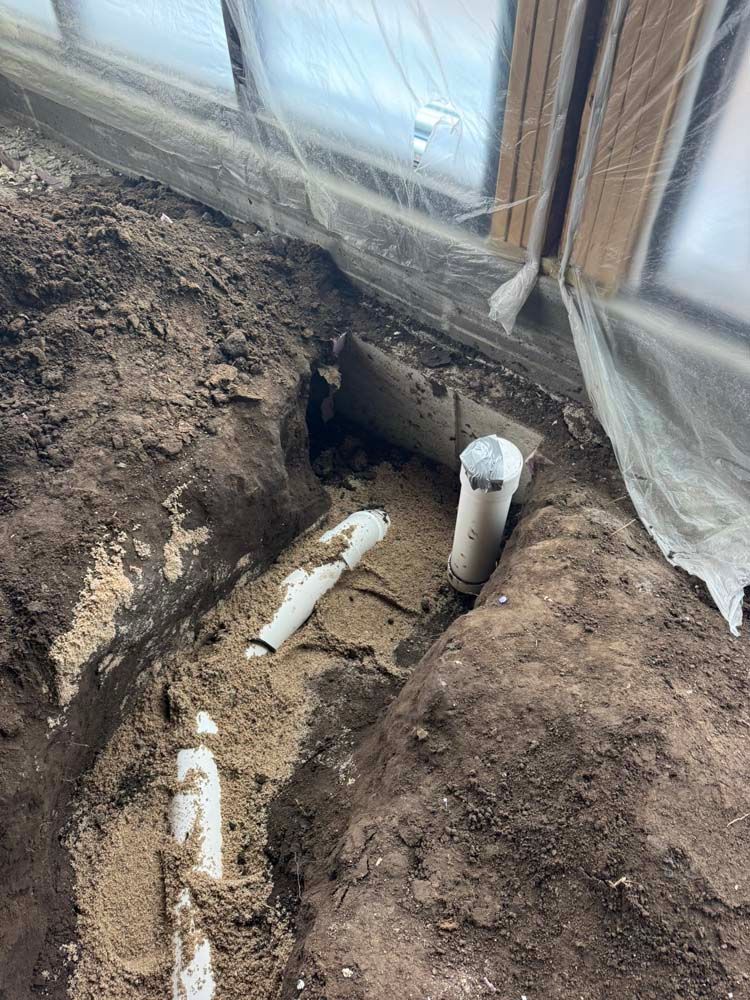Pipes in a trench beneath a plastic-covered structure, likely a greenhouse, in dirt.