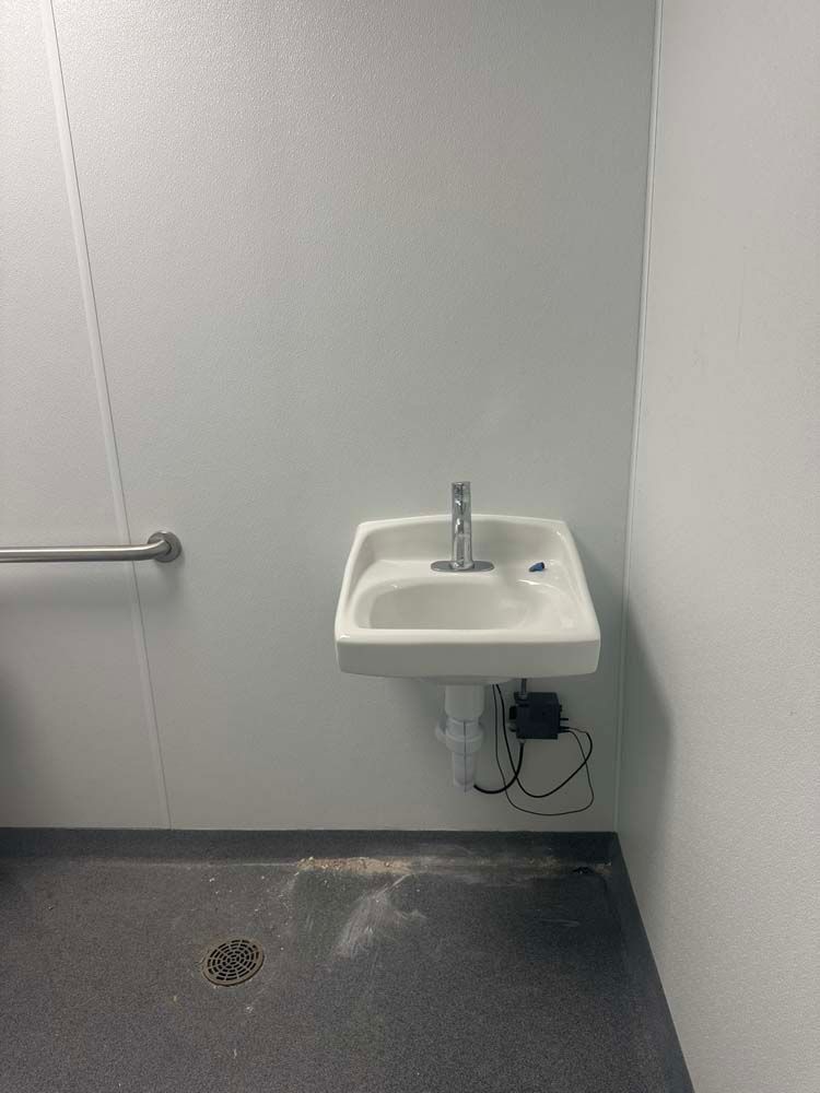 White wall-mounted sink in a plain room. Silver faucet and drain. A handrail is on the left.