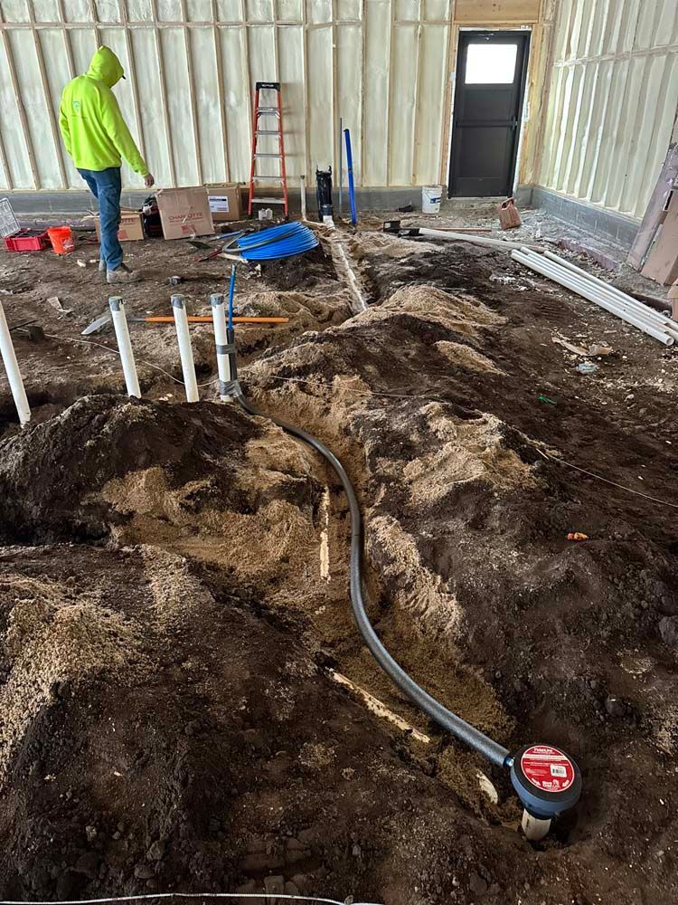 Construction site: Worker in yellow jacket, digging, exposed plumbing, dirt floor.