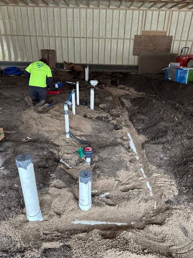 Plumbing installation in progress: a worker in a neon green shirt works amid pipes and sand on a construction site.