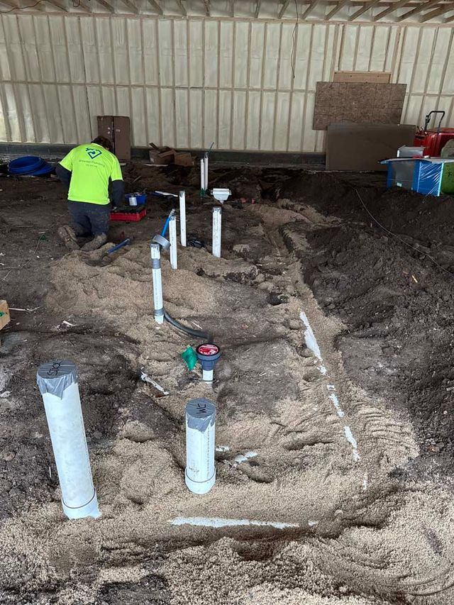 Plumbing installation in progress: a worker in a neon green shirt works amid pipes and sand on a construction site.