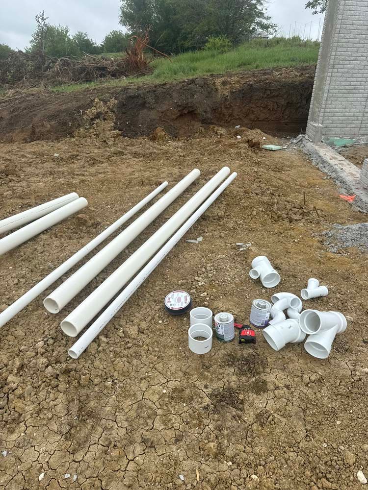 White PVC pipes and fittings on a dirt ground near a building under construction.