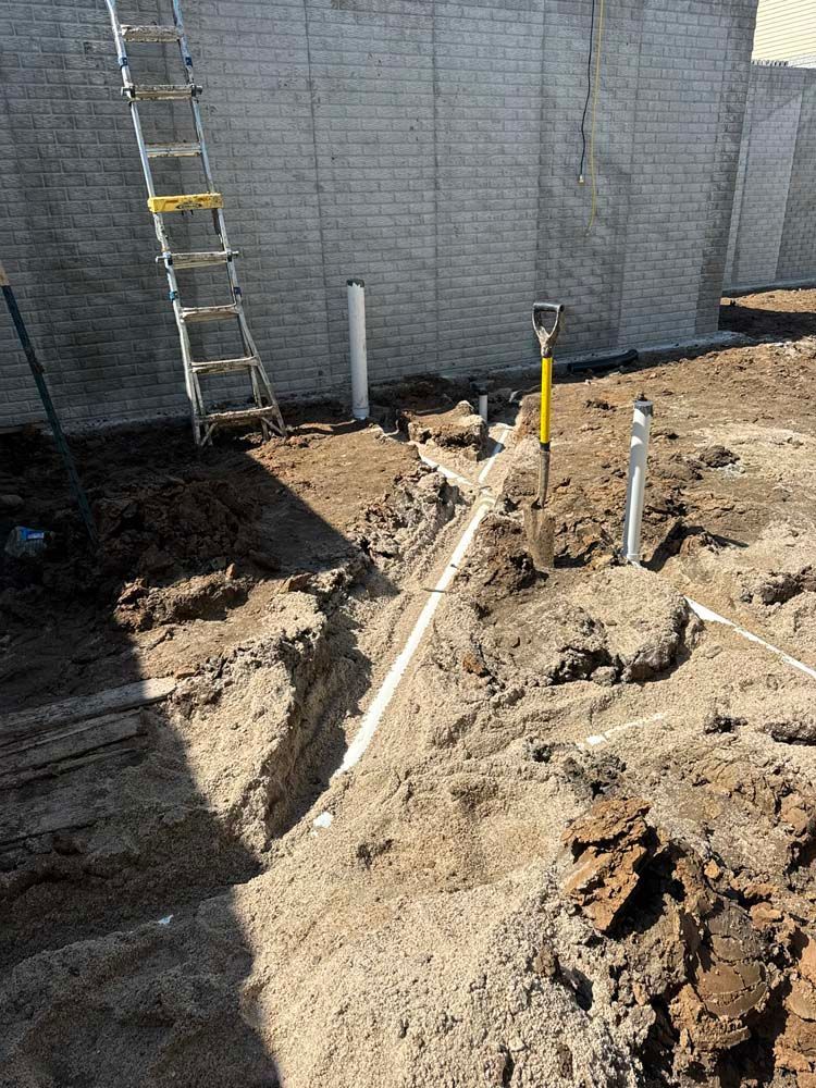 Construction site with a trench dug, white pipes, and a ladder leaning against a brick wall.