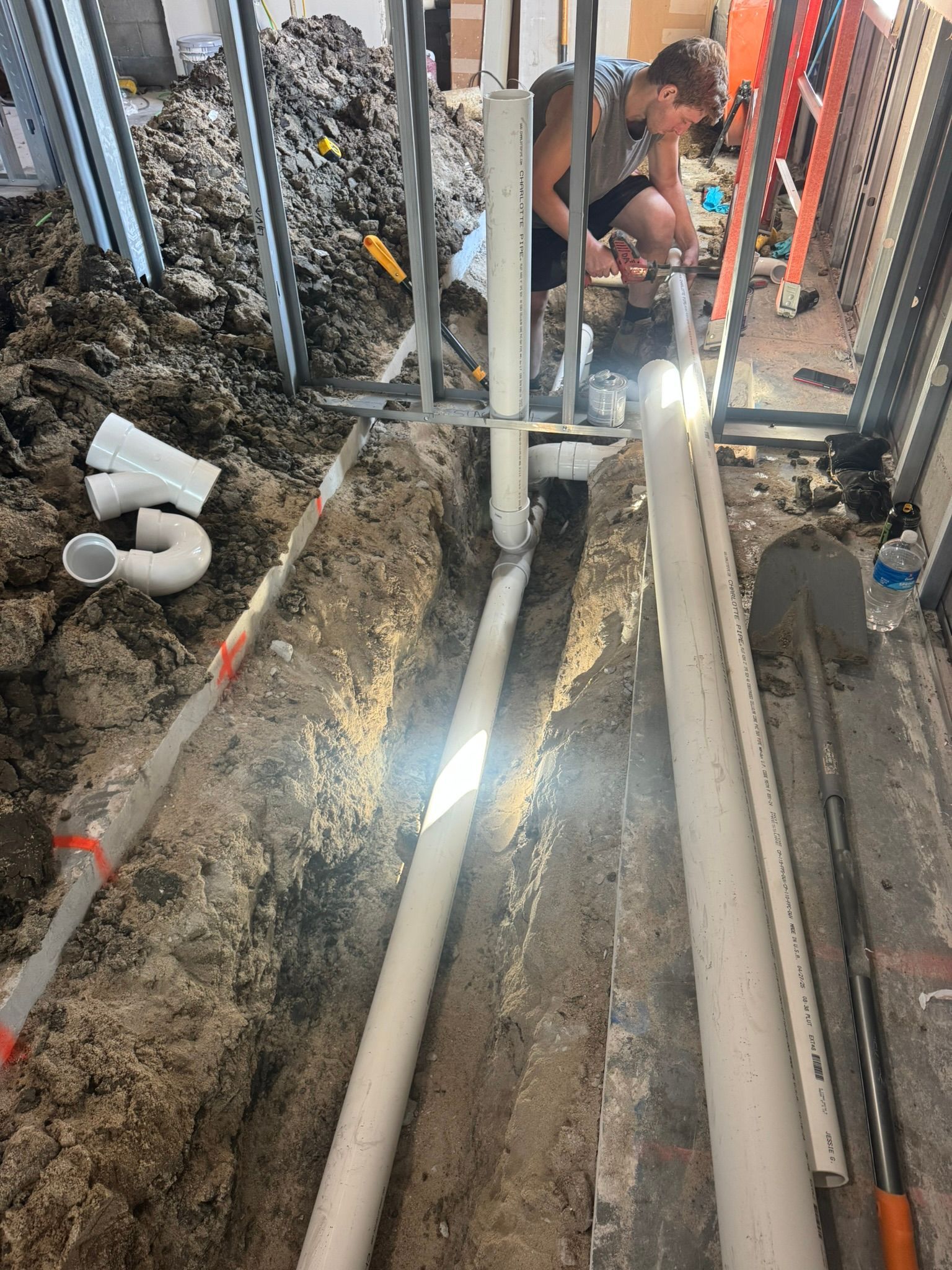 Plumbing work in progress: a person crouches over pipes in a trench. PVC pipes, dirt, tools, and metal framing are visible.