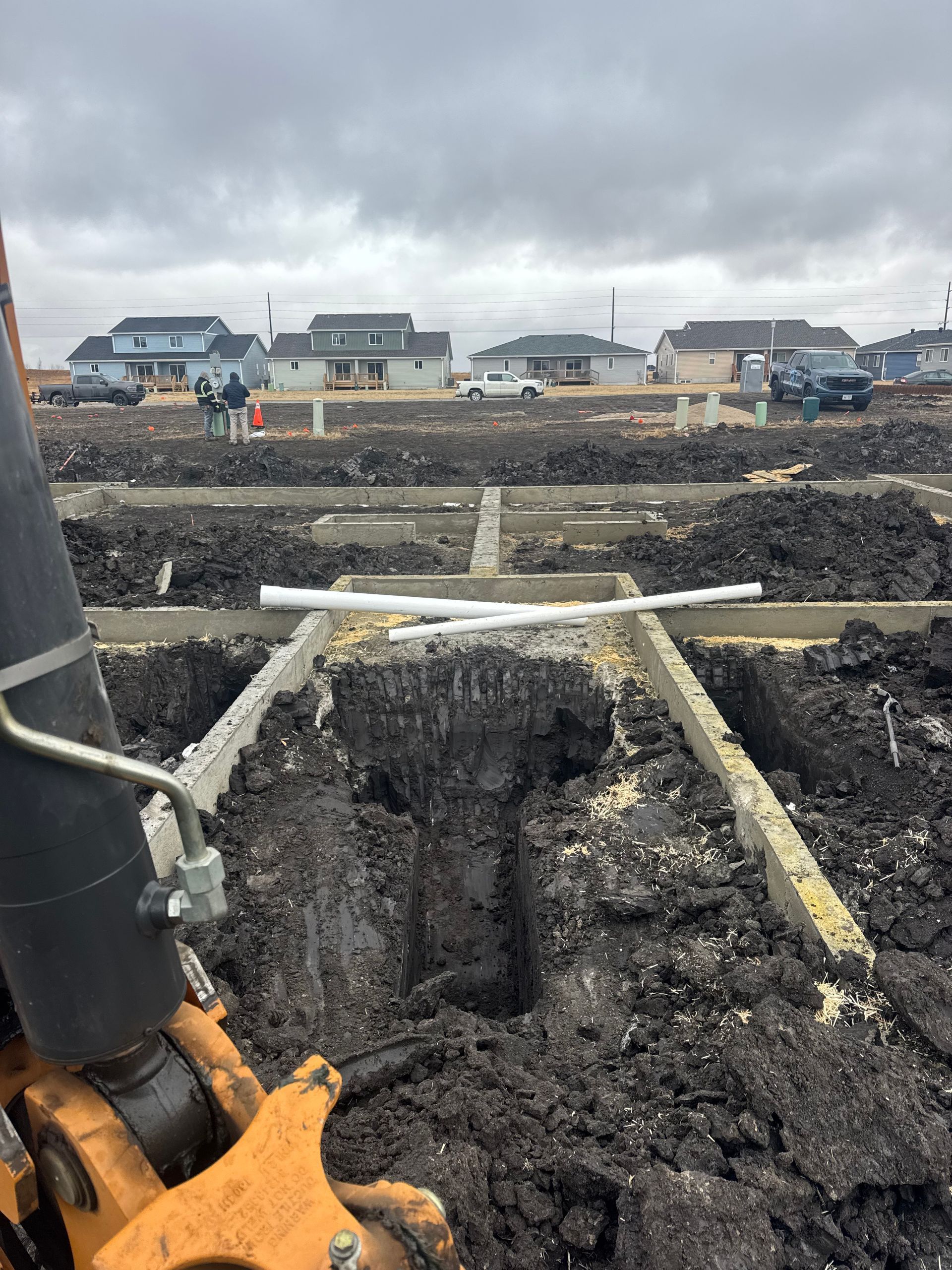 Excavation site with trenches for foundations, residential neighborhood in background. Overcast sky.