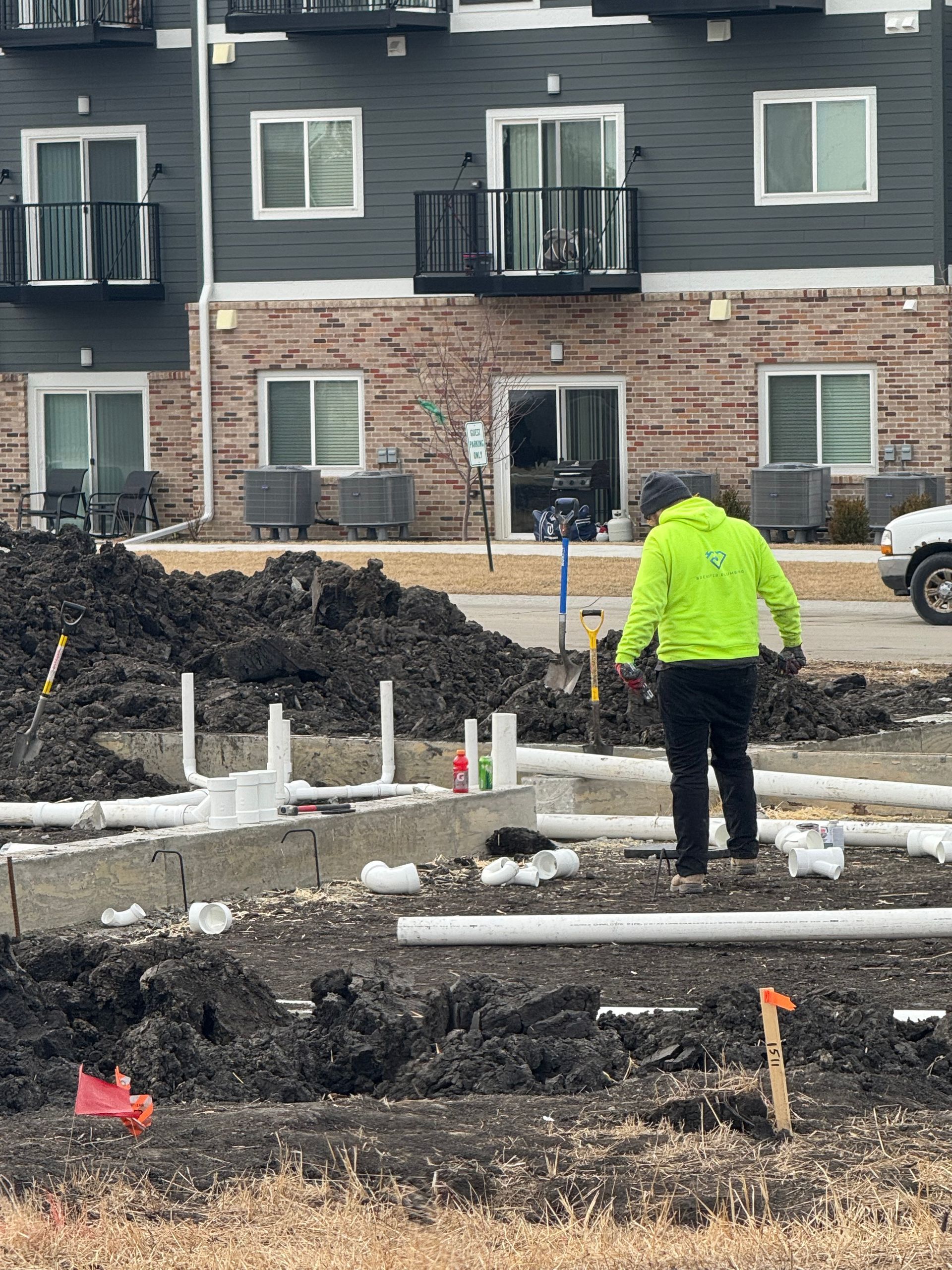 Construction worker in neon jacket surveying plumbing pipes near an apartment building.
