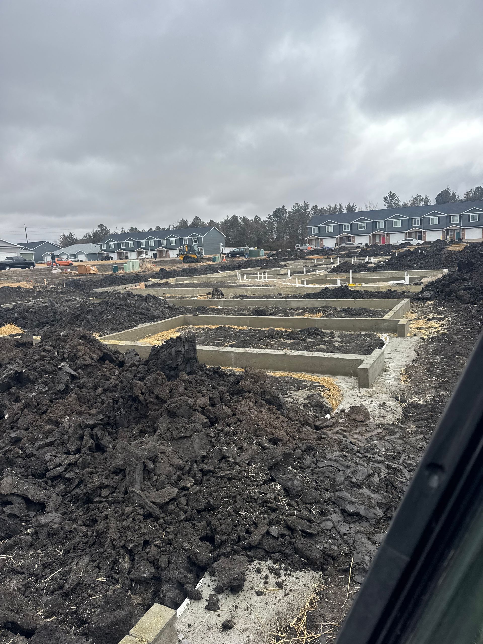 Construction site: foundations laid, dark soil, cloudy sky, distant buildings.