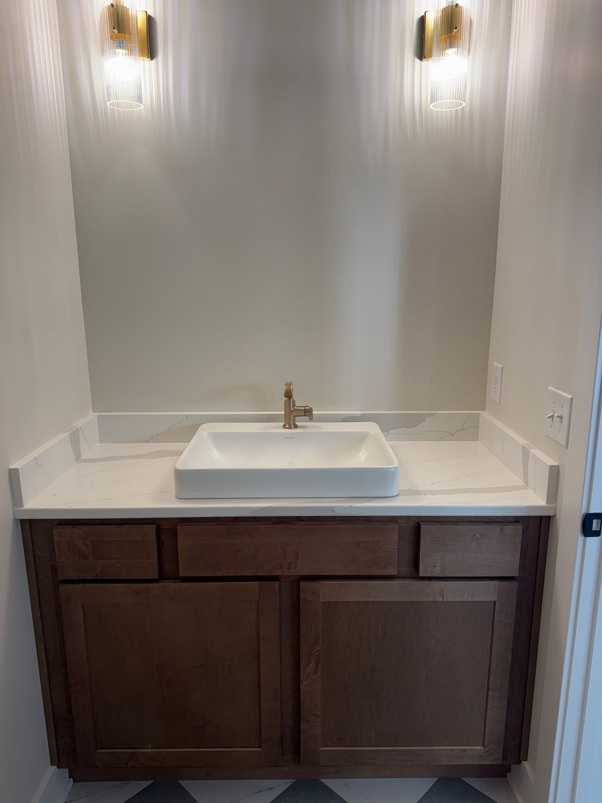 A powder room with a white sink, gold faucet, brown cabinet, and two gold sconces.