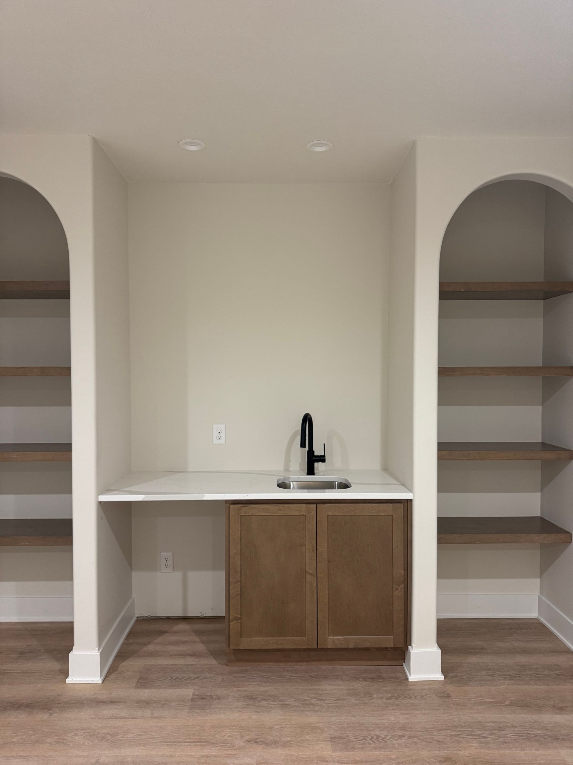 Built-in bar with arched shelves, a sink, and cabinets, in a neutral-toned room with wood-look flooring.