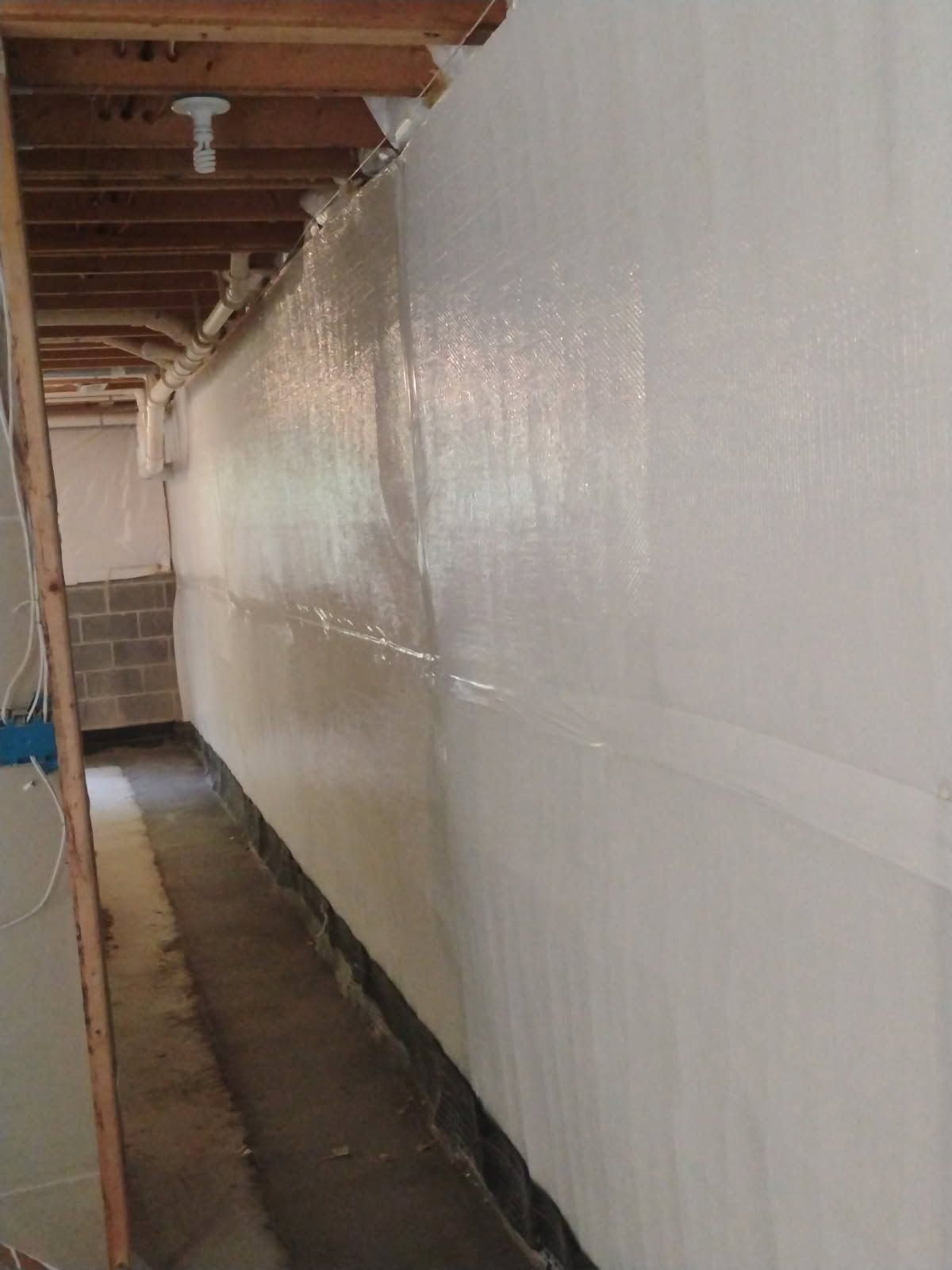 Narrow unfinished basement hallway with exposed beams and a long white plastic-covered wall