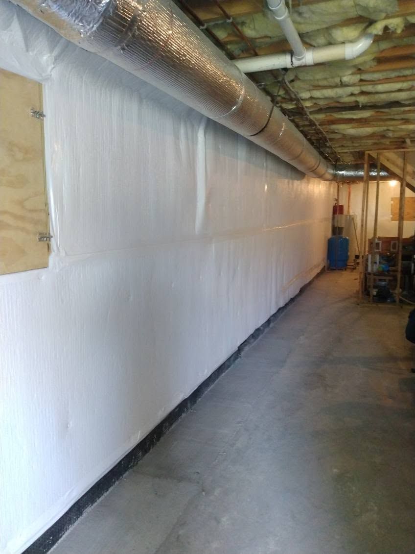Long white insulated wall in a basement or crawlspace with exposed pipes and concrete floor.