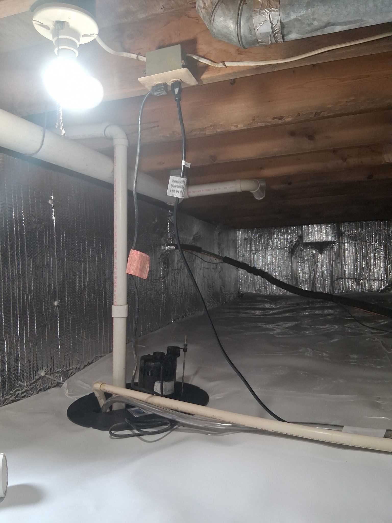 Basement crawlspace with exposed pipes, wiring, and a single bare bulb illuminating insulation.