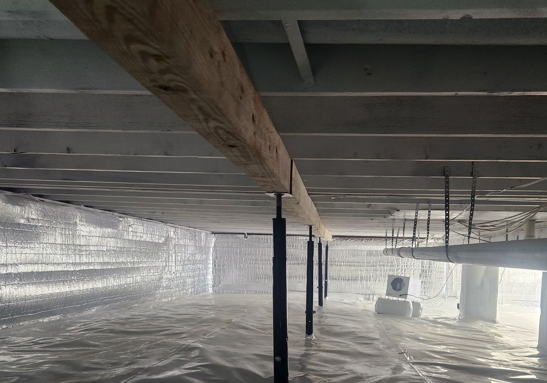 Plastic-covered crawlspace with exposed wooden beams and utility pipes under a house