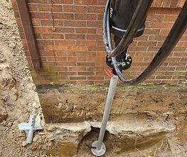 Drill rig with auger boring into soil beside a brick wall foundation, with a measuring rod nearby