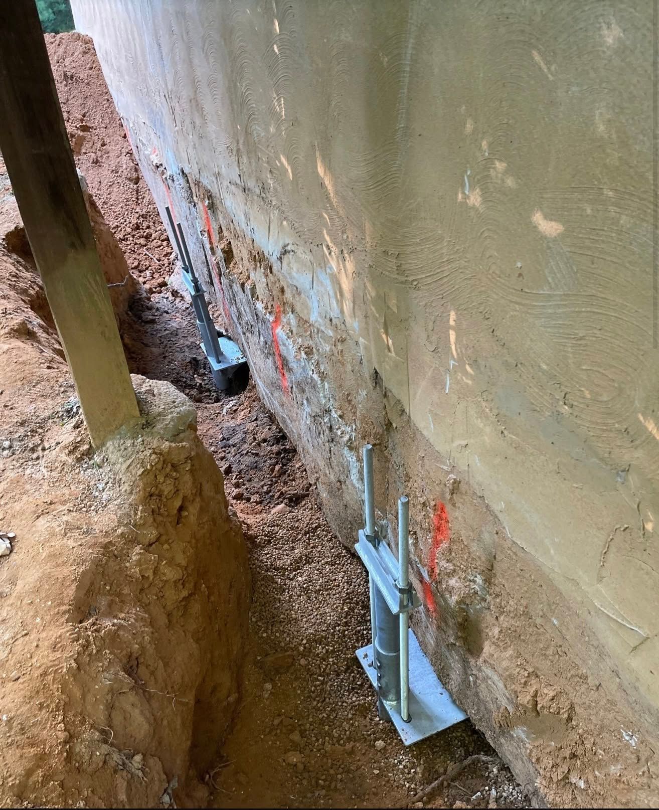 Foundation wall with anchor bolts and brackets in a narrow dirt trench beside a concrete structure.