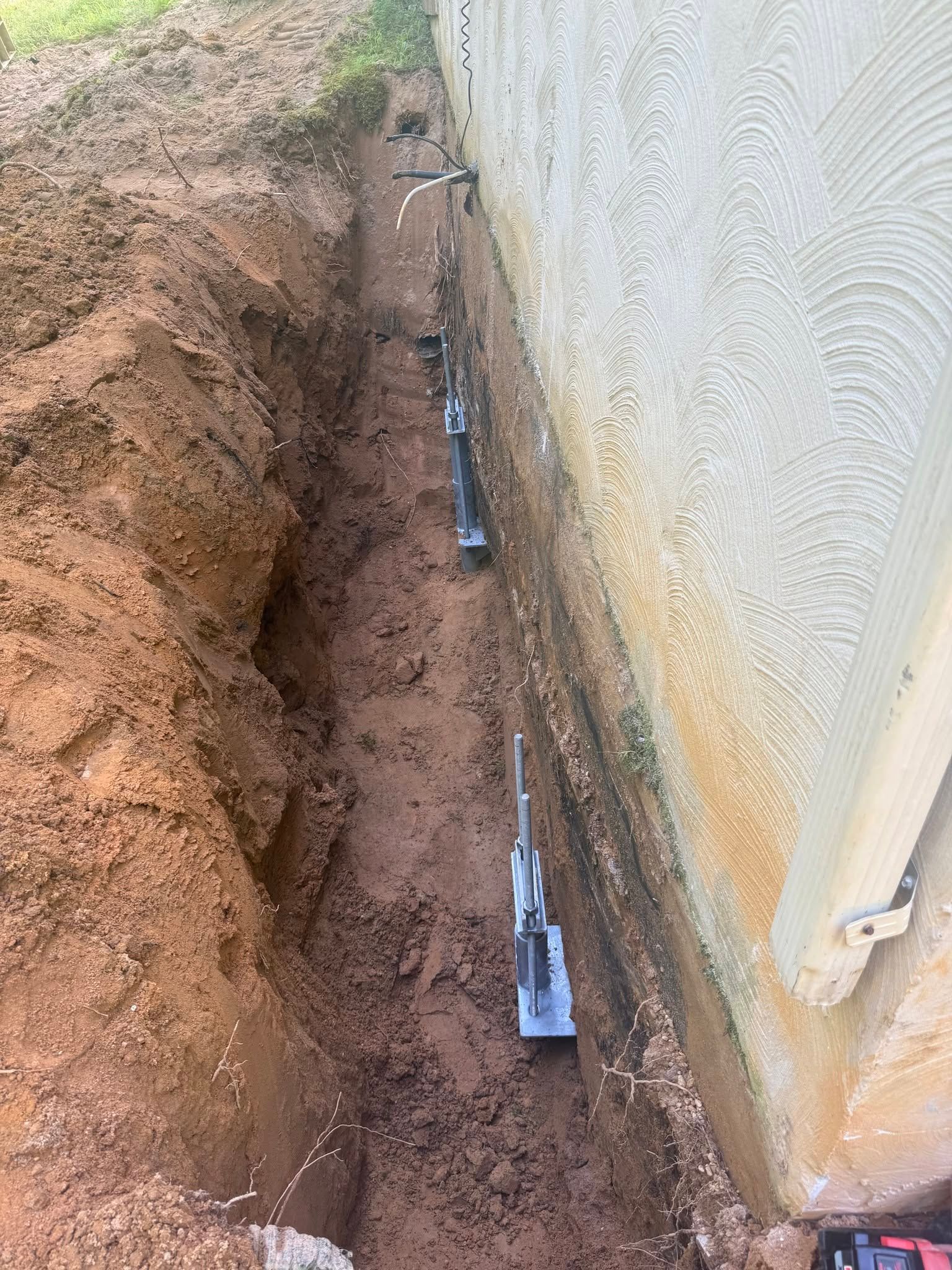 Trench beside a concrete wall, with pipes and fittings laid along the bottom in muddy soil.