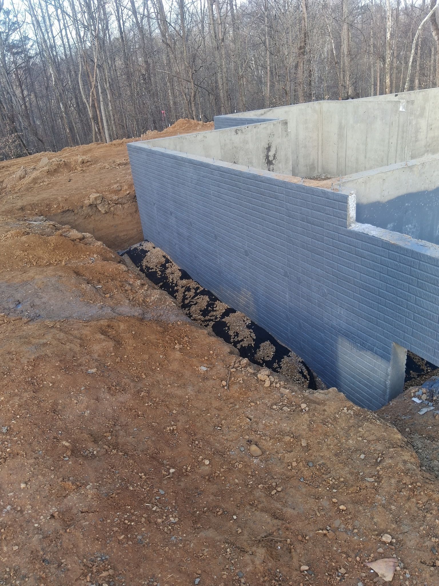 Foundation walls under construction with blue waterproofing, dirt excavation, and a wooded background.