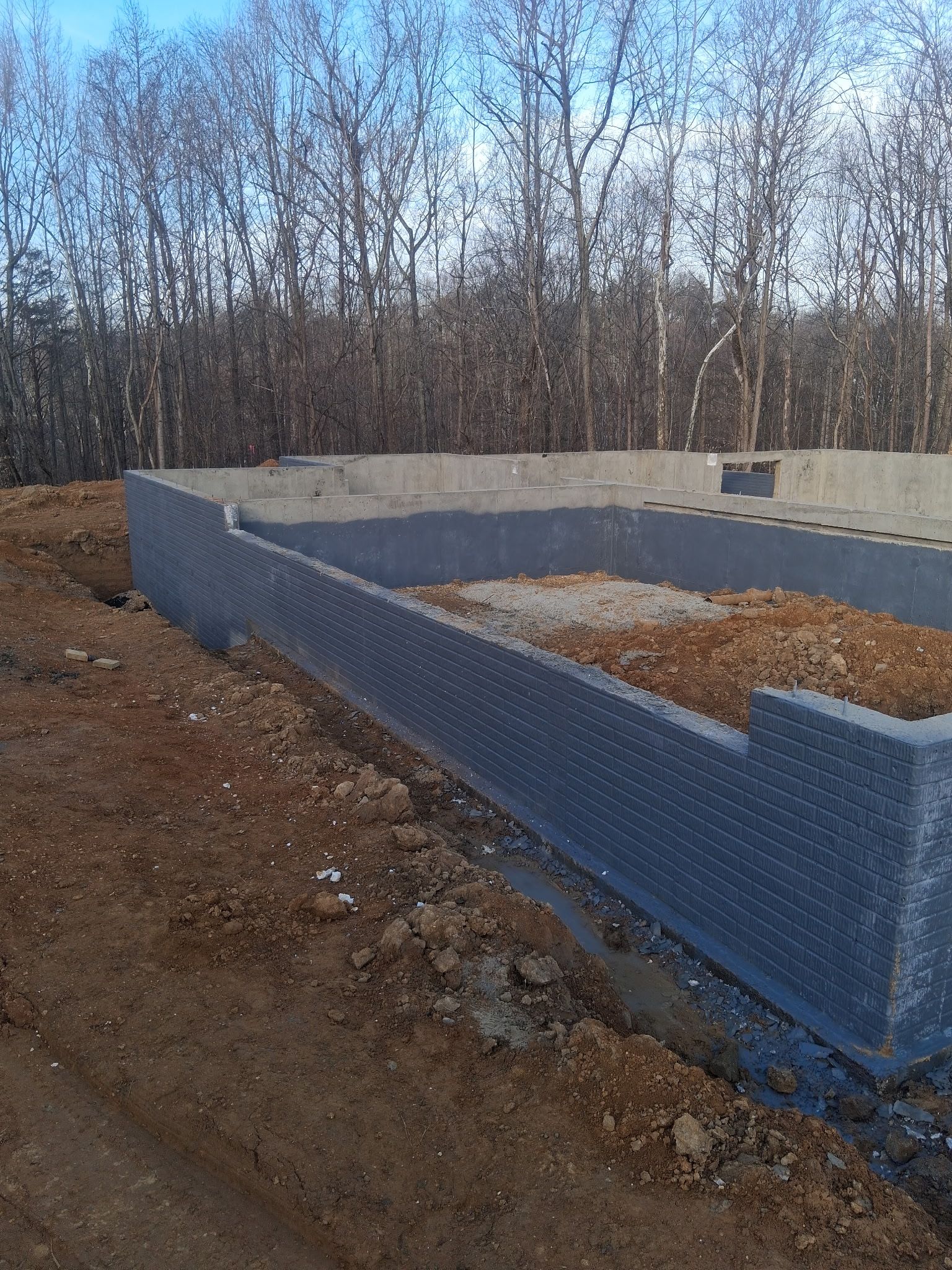 Concrete foundation walls at a construction site beside a dirt embankment and leafless trees.