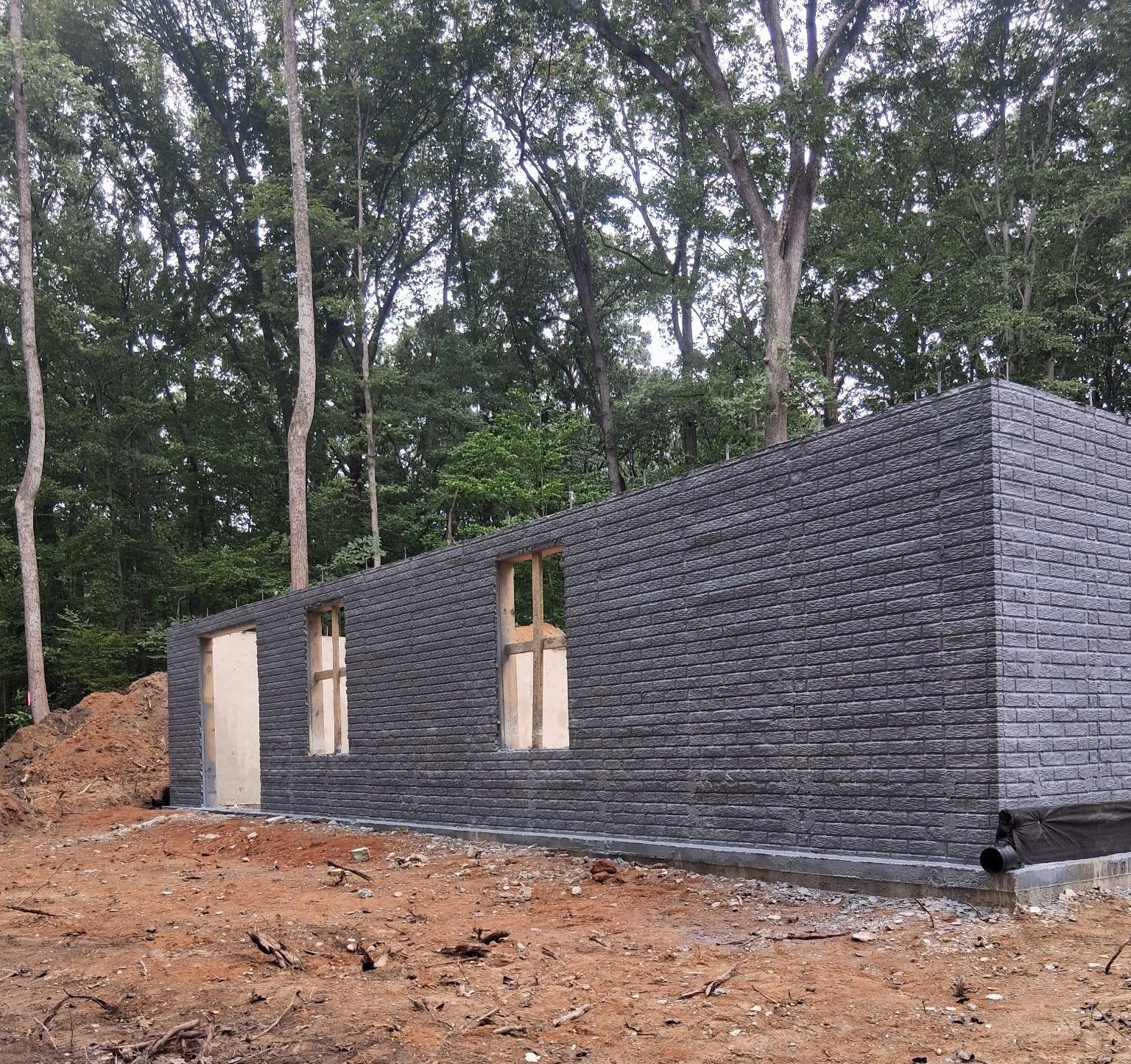 Stone-clad house under construction in a wooded area, with unfinished window openings and exposed foundation.