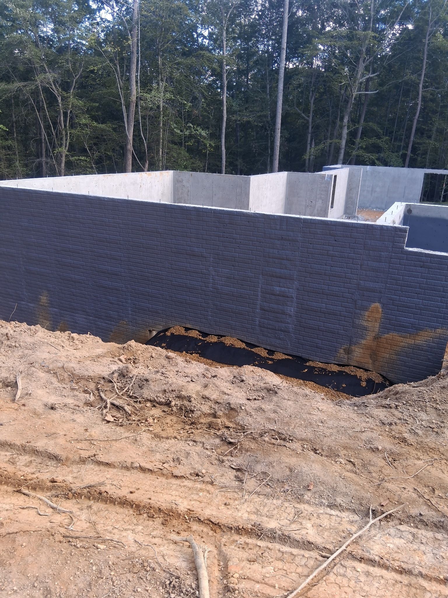 Partially built concrete wall with black mesh barrier on a dirt construction site, trees in background