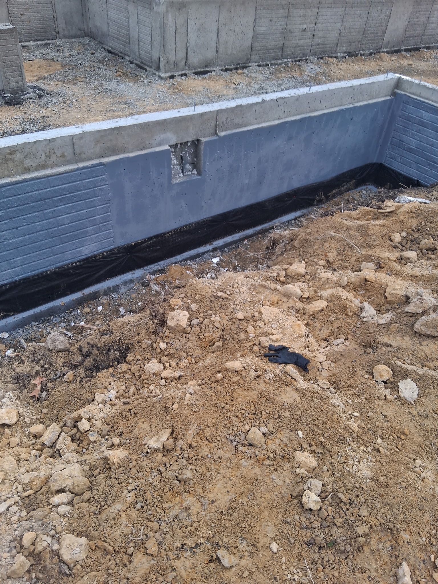 Foundation wall with black waterproofing, drainage strip, and dirt in the foreground at a construction site
