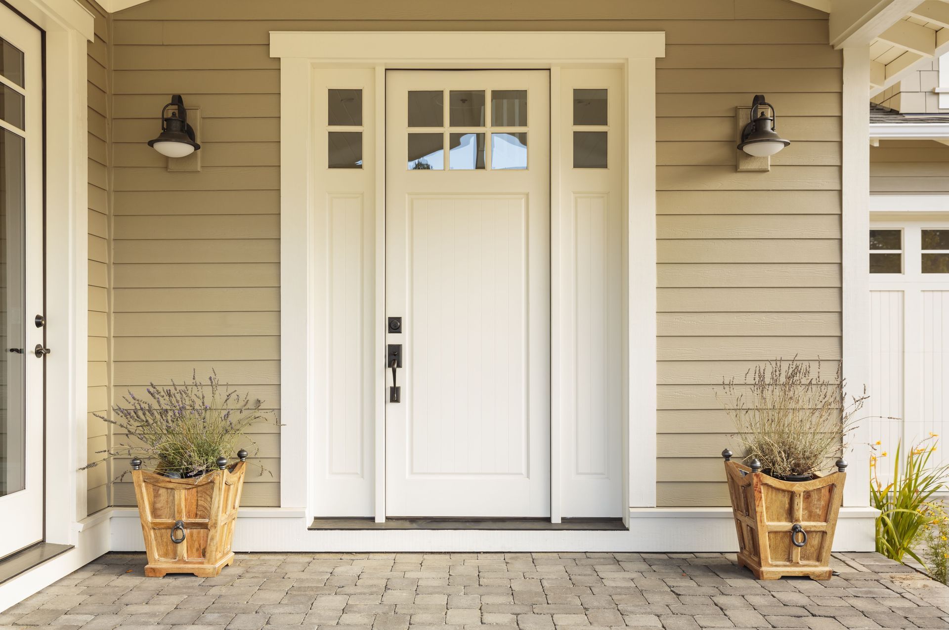 White front door with sidelights and transom, flanked by potted plants, on a beige siding house.