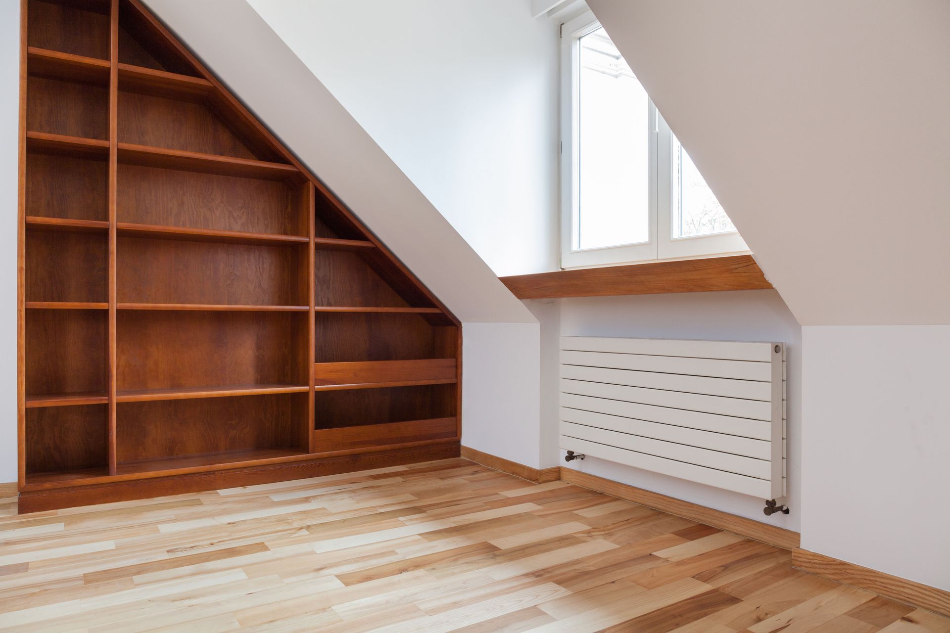 Wooden bookshelf built into an attic wall, beside a window and radiator; hardwood floor.