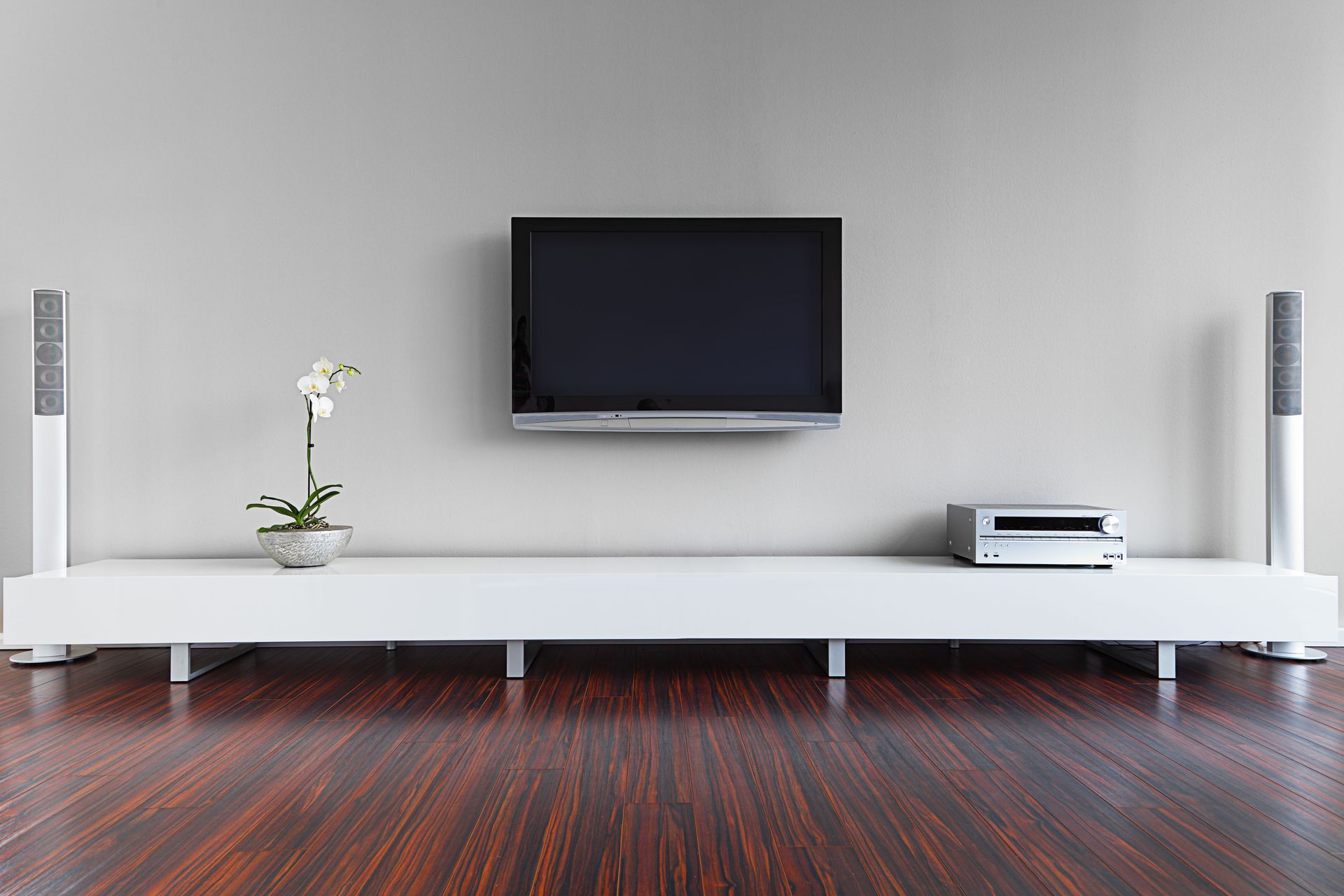 A TV is mounted above a white entertainment console, flanked by speakers, with an orchid.