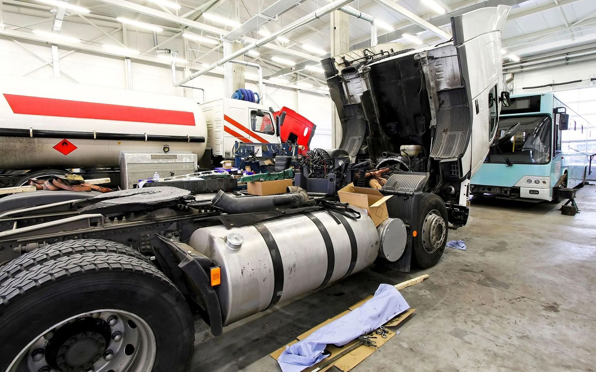 A mechanic shop interior with a truck cab tilted forward for engine repair, a tanker trailer, and a bus in the background.