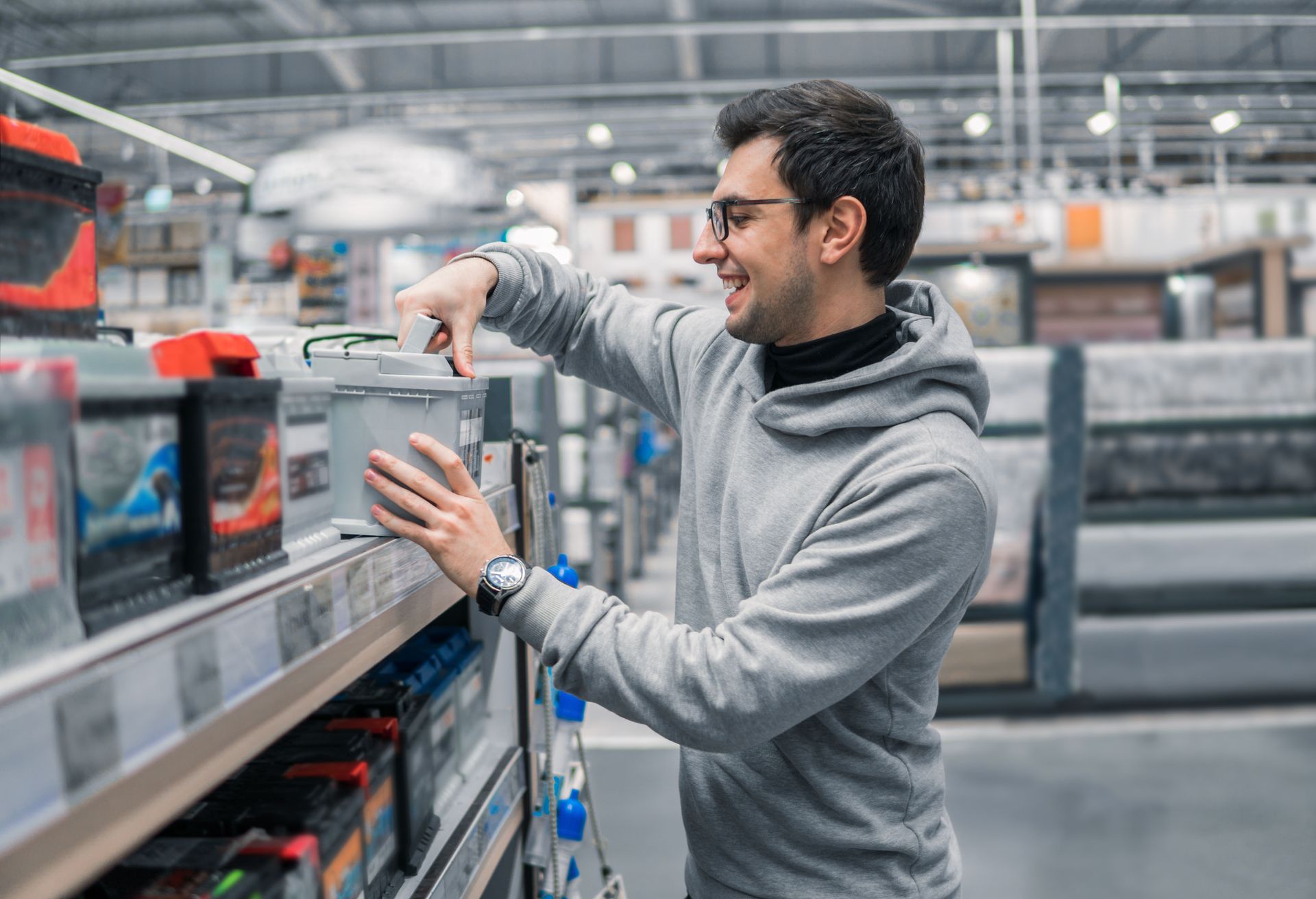 A person wearing a grey hoodie and glasses smiles while selecting a car battery from a shelf in an automotive store.