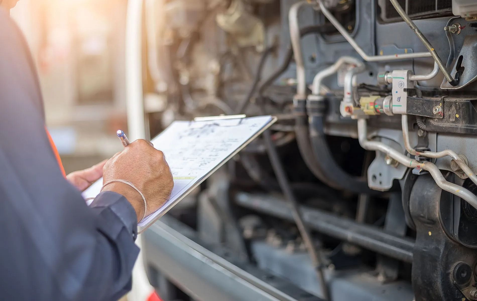 A technician in a uniform writes on a clipboard while inspecting the exposed engine components of a vehicle.