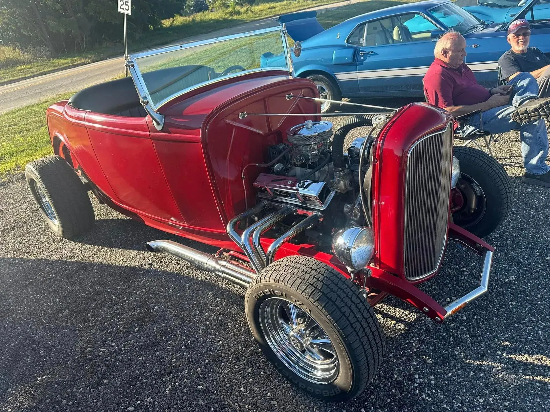 A bright red, open-top hot rod parked on gravel at a car show, with two people sitting in a blue car behind it.