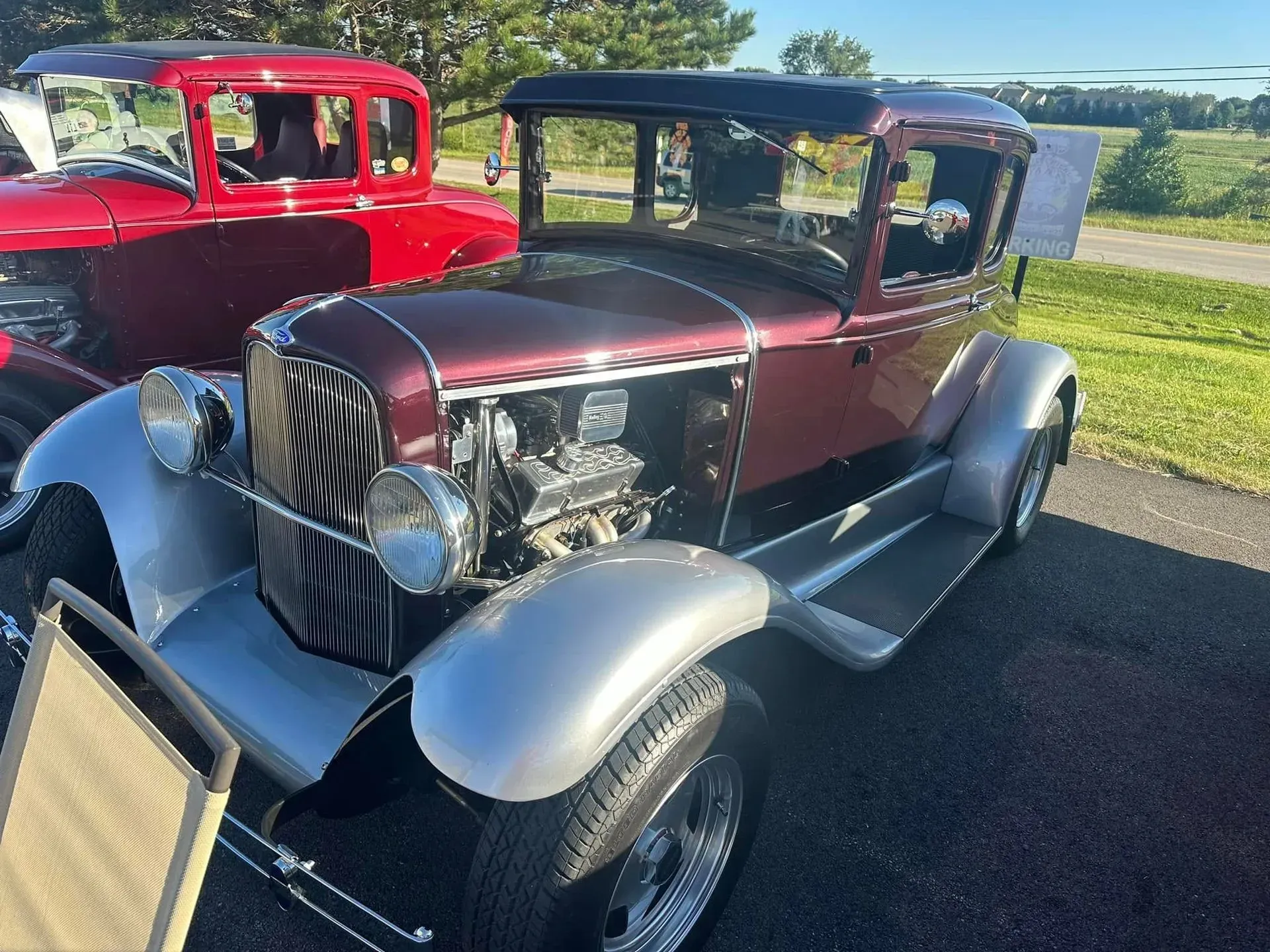 A maroon and silver vintage hot rod parked on asphalt, with a partially visible red classic car in the background.