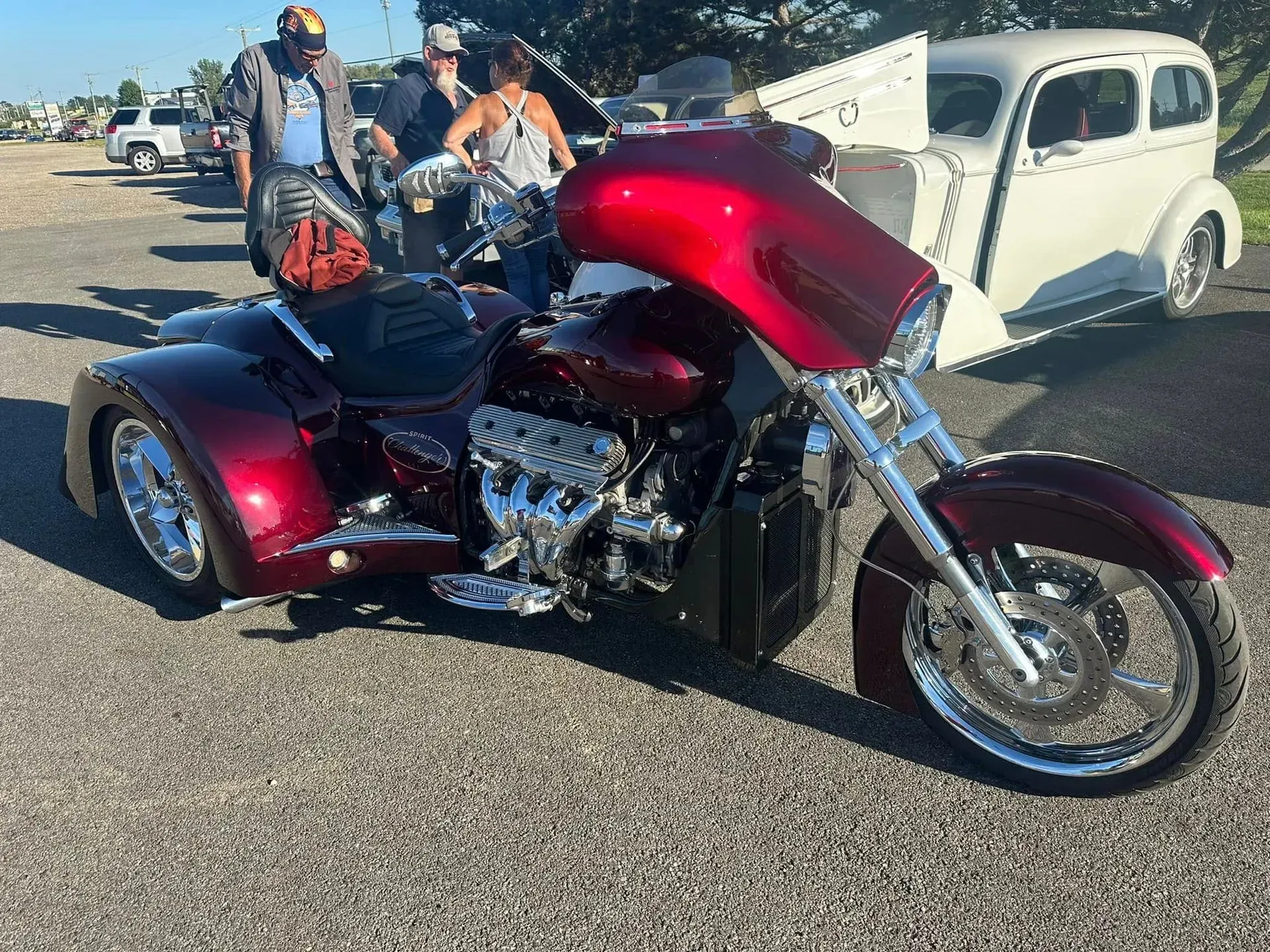 A custom deep red three-wheeled motorcycle parked on gravel, with people and a white vintage car in the background.