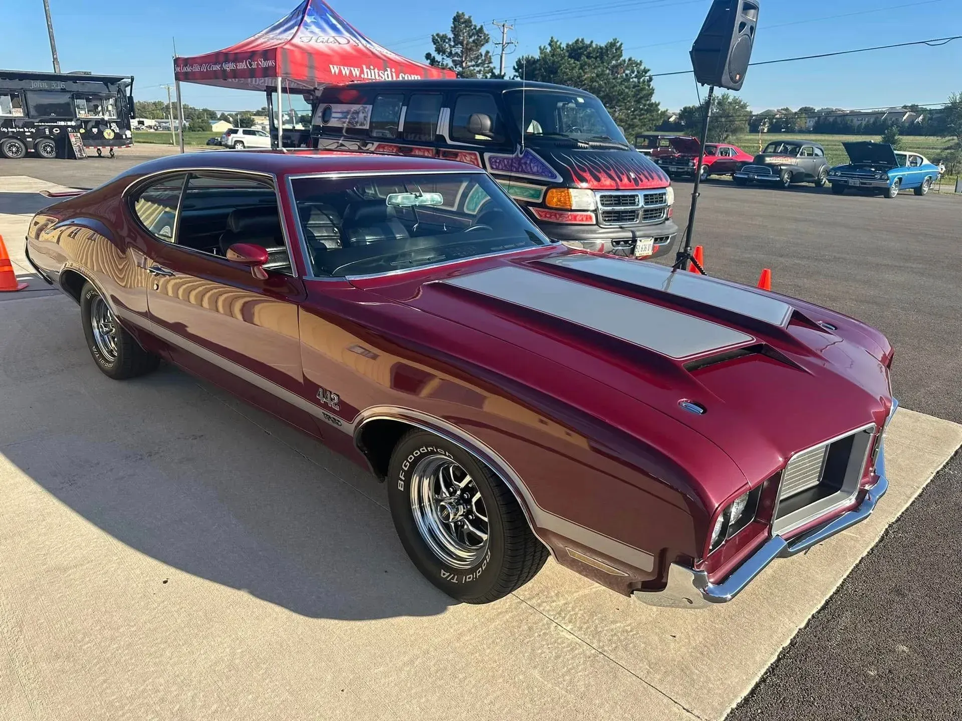 A glossy burgundy muscle car with silver hood stripes parked on pavement at an outdoor car show.