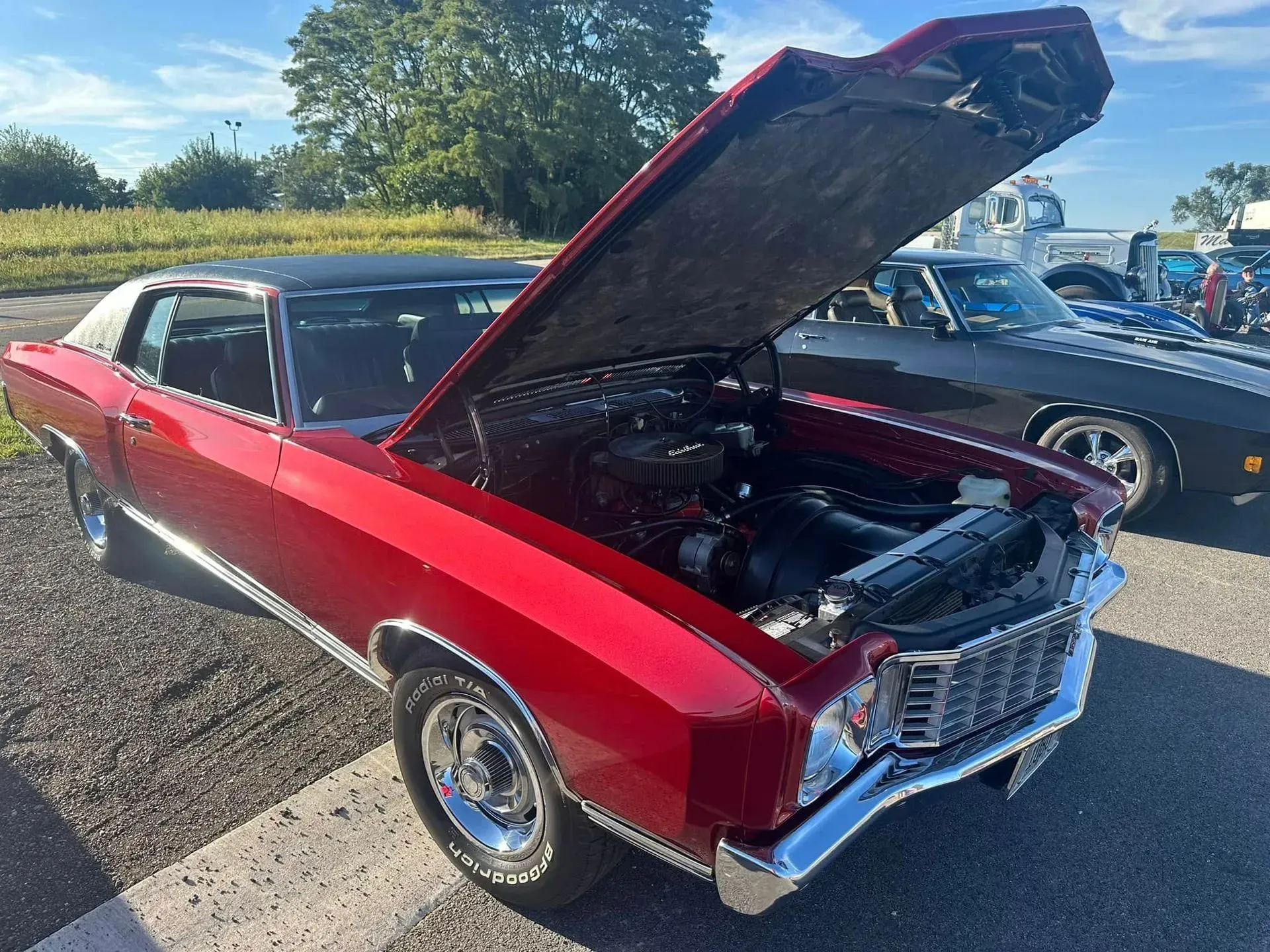 A cherry-red vintage Chevrolet Monte Carlo with its hood open, parked in an outdoor lot on a sunny day.