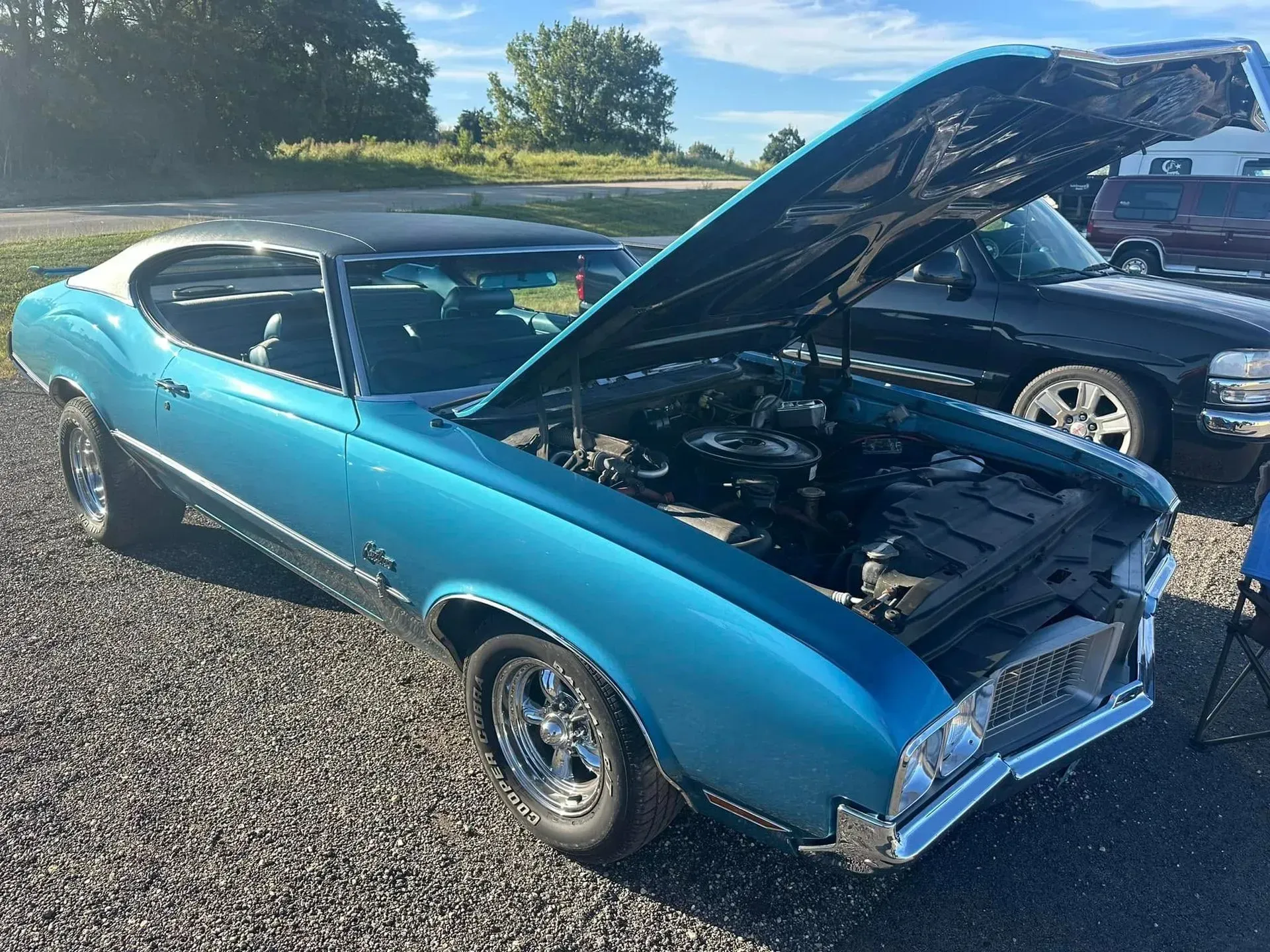 A bright metallic blue vintage Oldsmobile Cutlass with its hood open, parked on a gravel lot under a sunny sky.