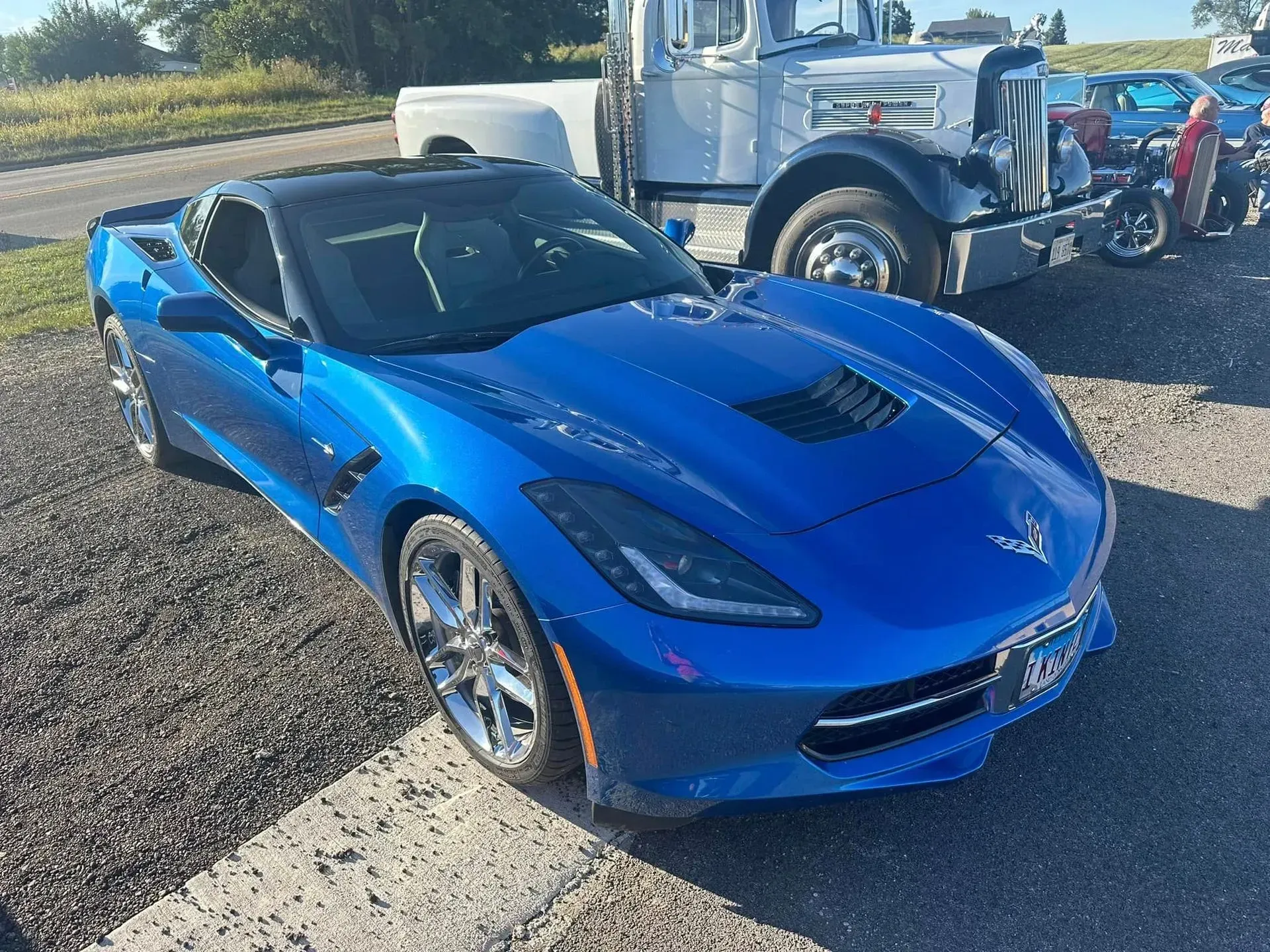 A bright blue Chevrolet Corvette parked on a gravel lot next to a white vintage truck.