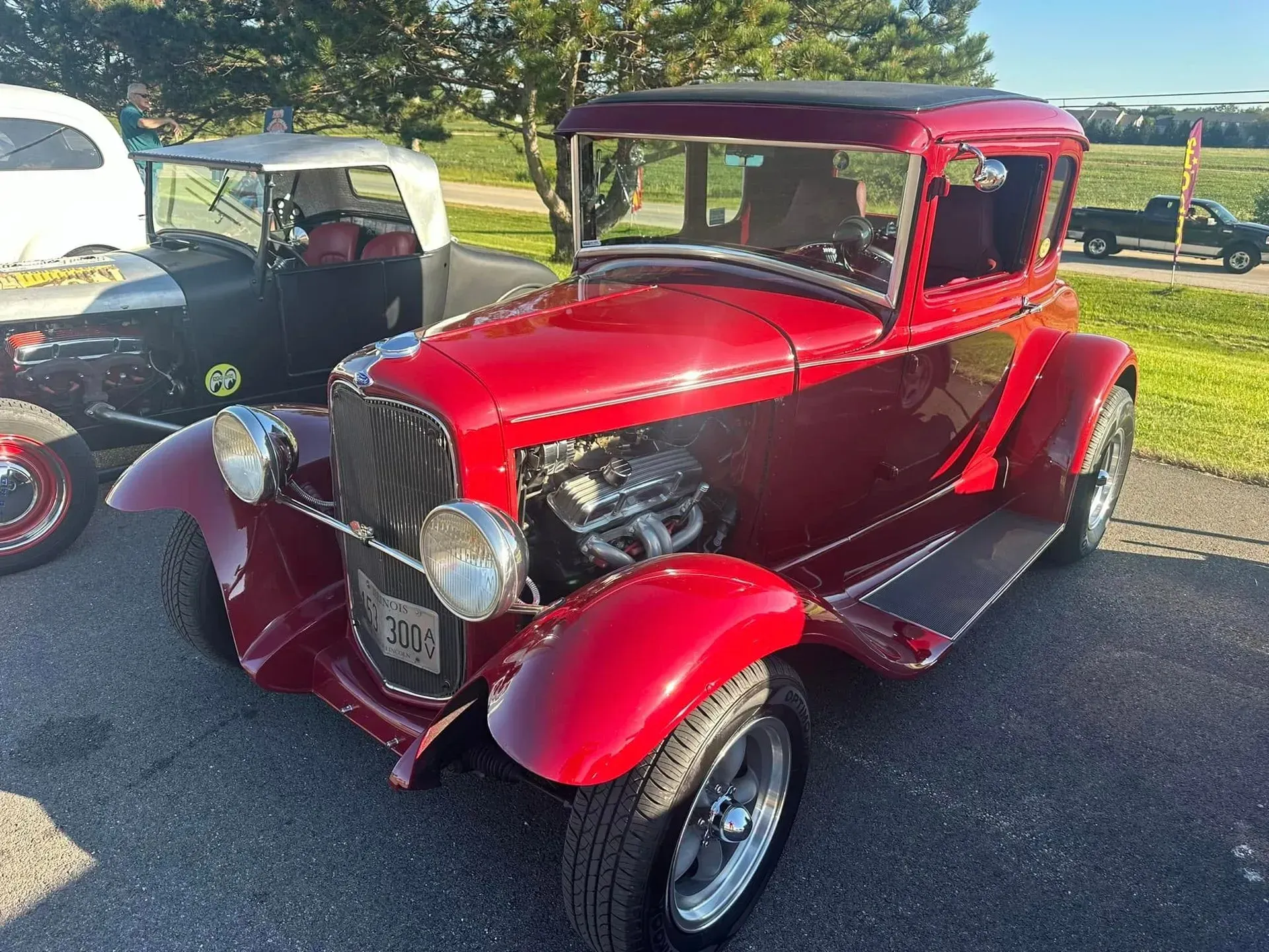 A bright red, vintage-style hot rod car parked outdoors on an asphalt lot, with another car partially visible behind it.